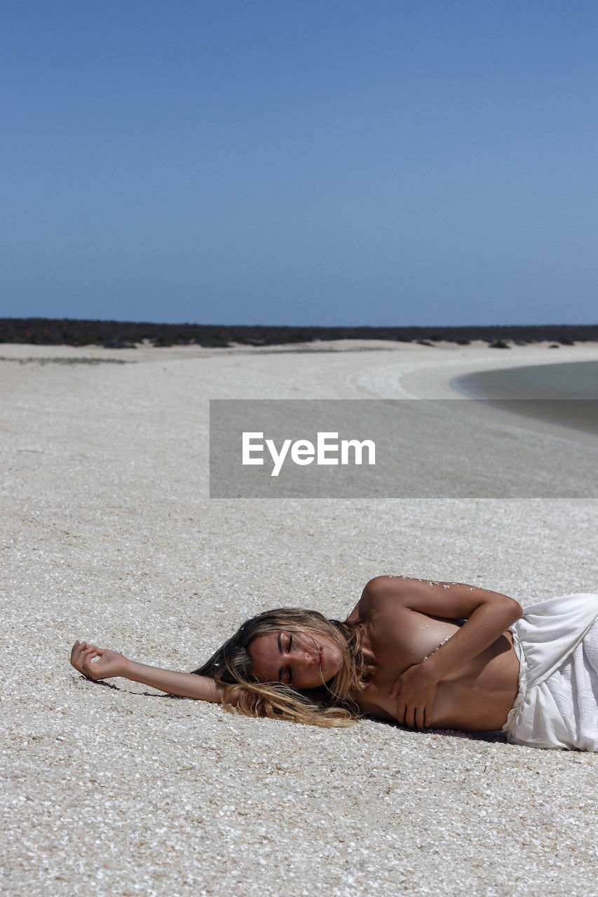Young blonde woman in white dress lying on the floor due to heat stroke on paradisiacal shell beach