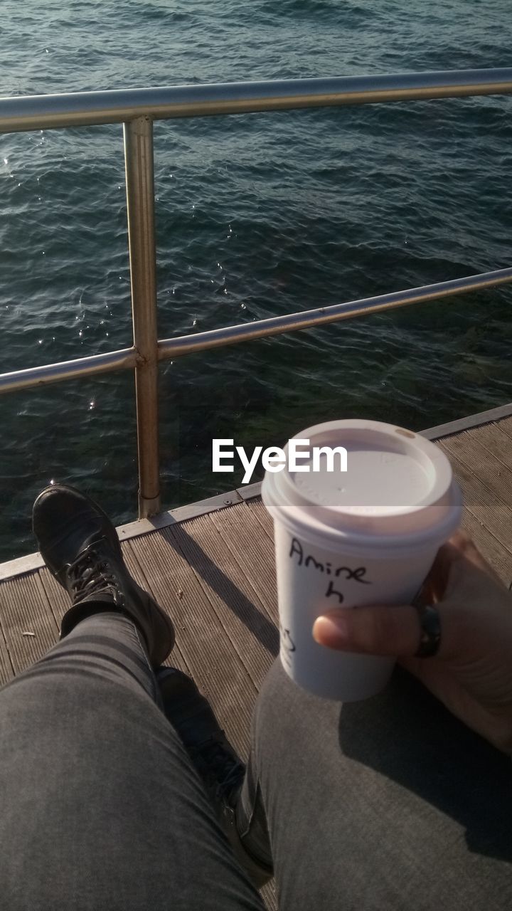 LOW SECTION OF PERSON WITH COFFEE CUP BY SEA