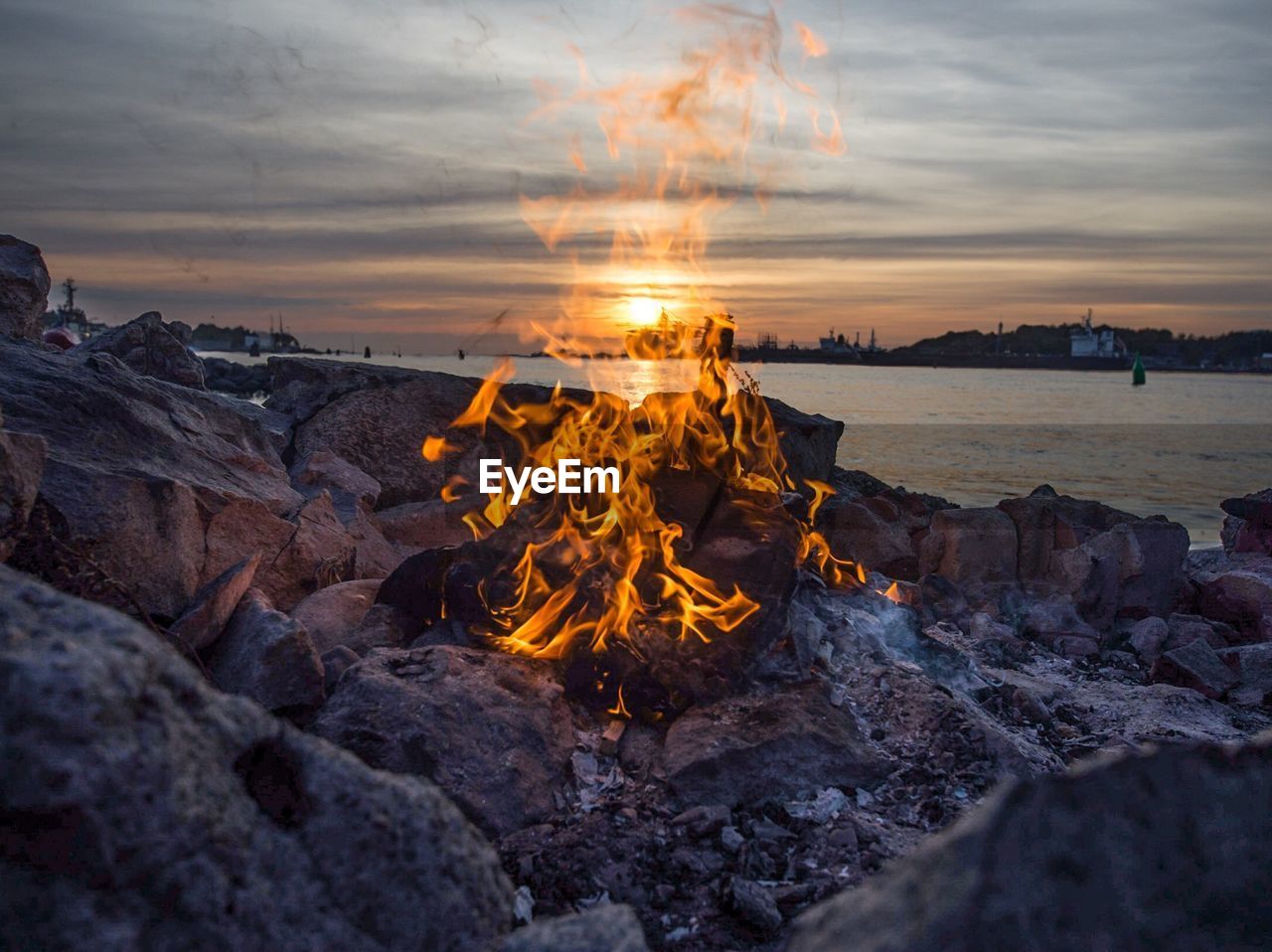 Close-up of campfire on rocks at lakeshore against cloudy sky during sunset