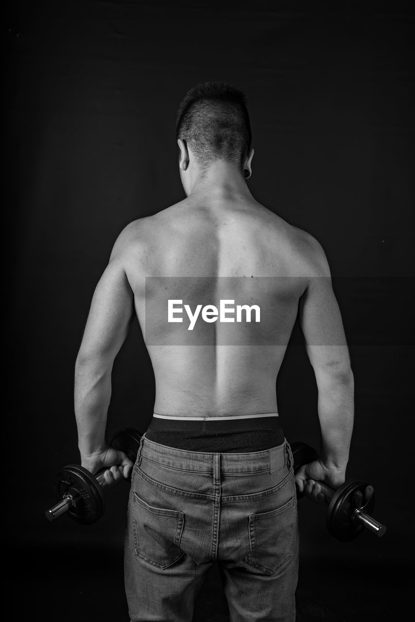 Rear view of shirtless man lifting dumbbells against black background