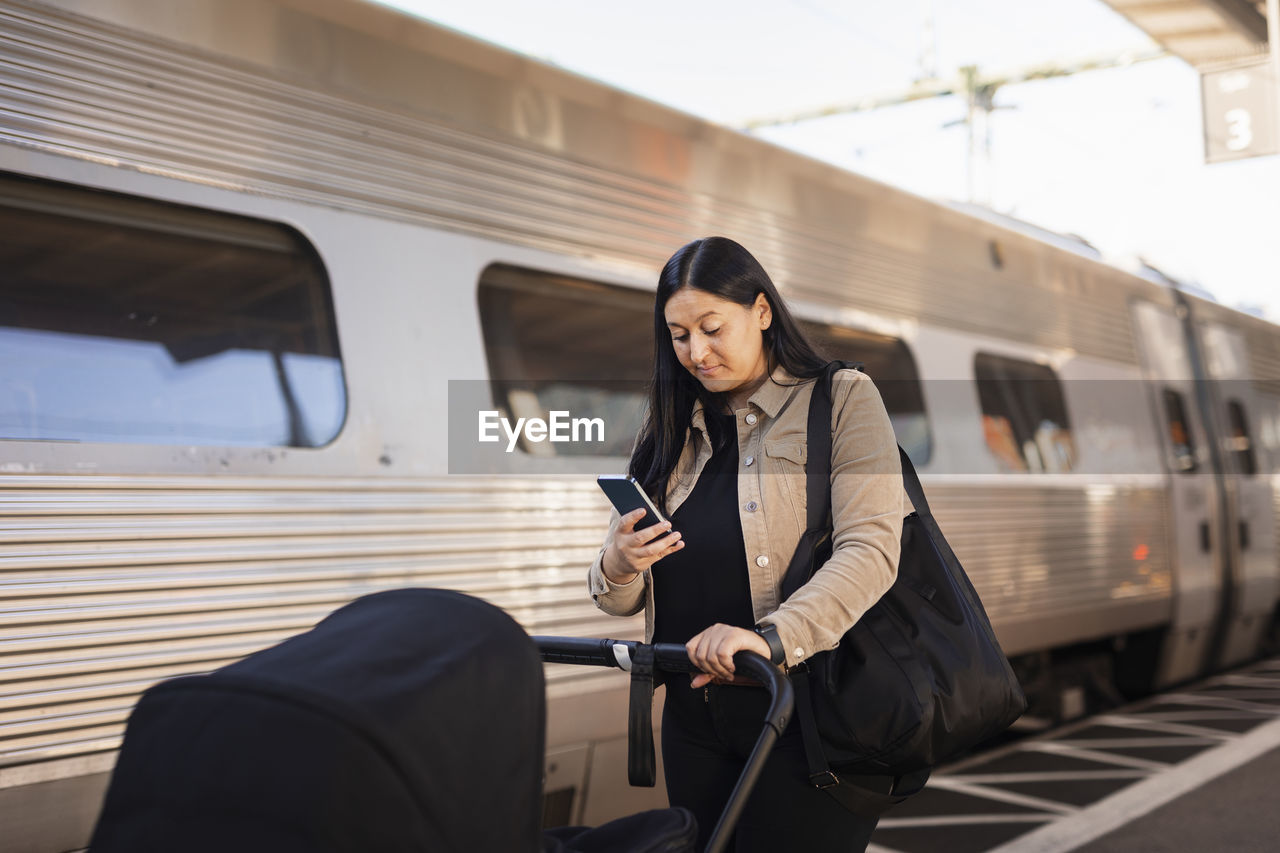 Mid adult woman at train station pushing pram and using cell phone