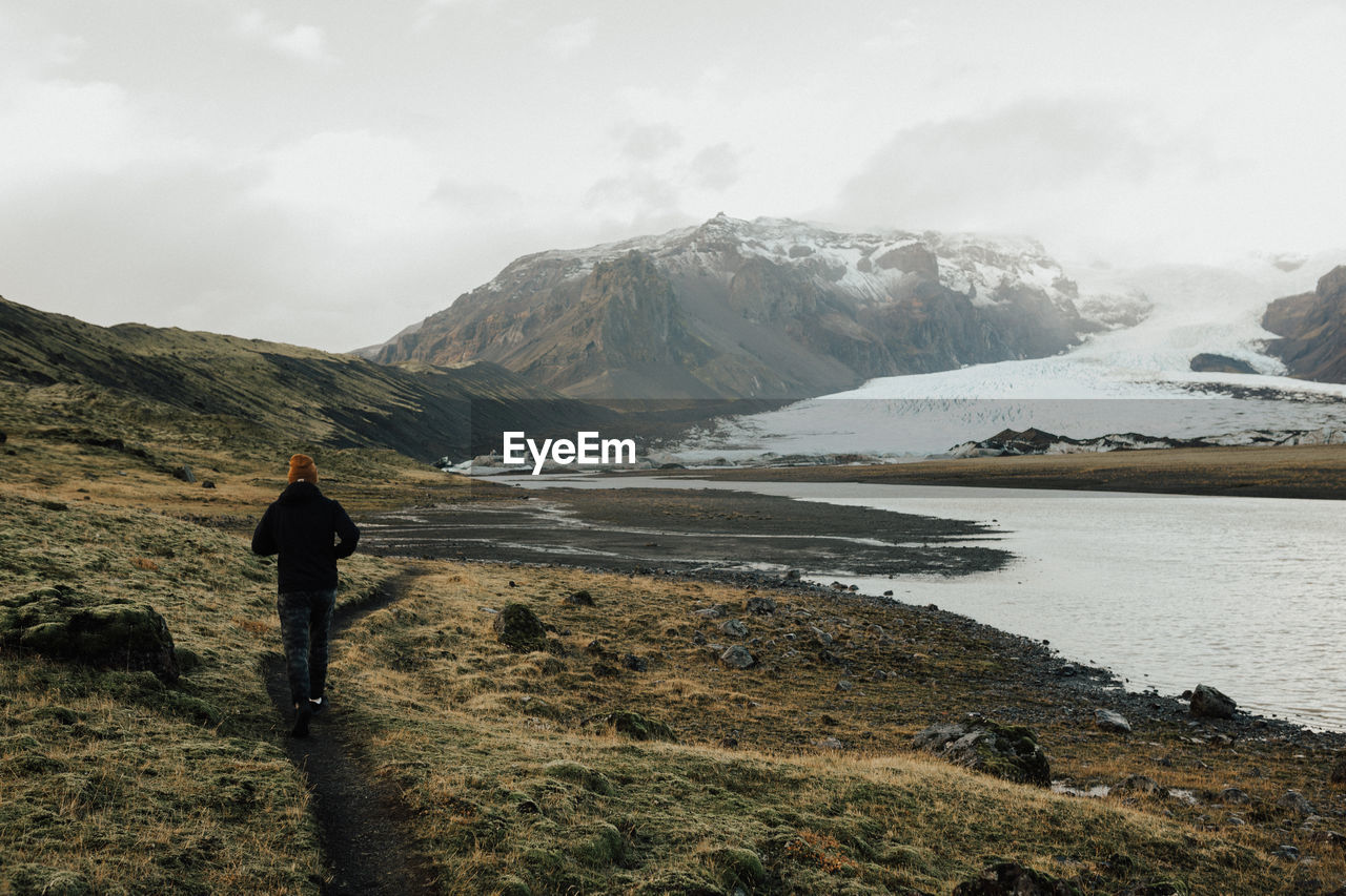 Rear view of man walking on landscape by lake against sky