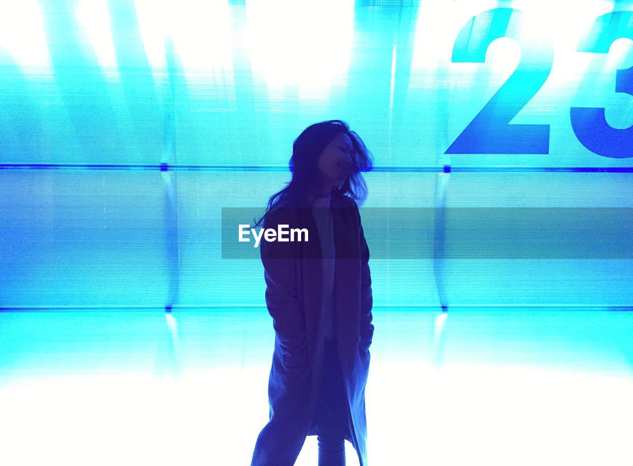 YOUNG WOMAN STANDING AGAINST ILLUMINATED BLUE WALL