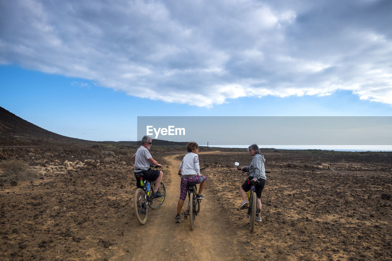 Friends with bicycles on dirt road against cloudy sky