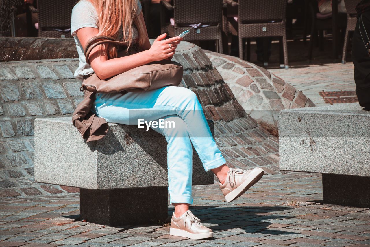 Low section of woman using mobile phone while sitting on retaining wall during sunny day