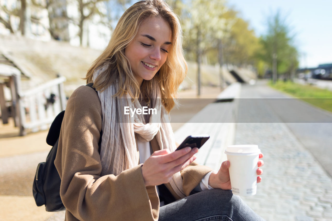 Woman with coffee cup using mobile phone in city