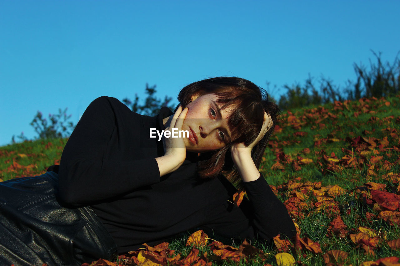 Portrait of young woman lying down on field against clear blue sky