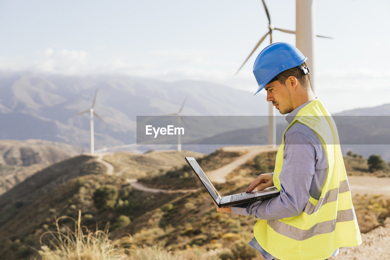 Engineer using laptop in front of wind turbines on sunny day