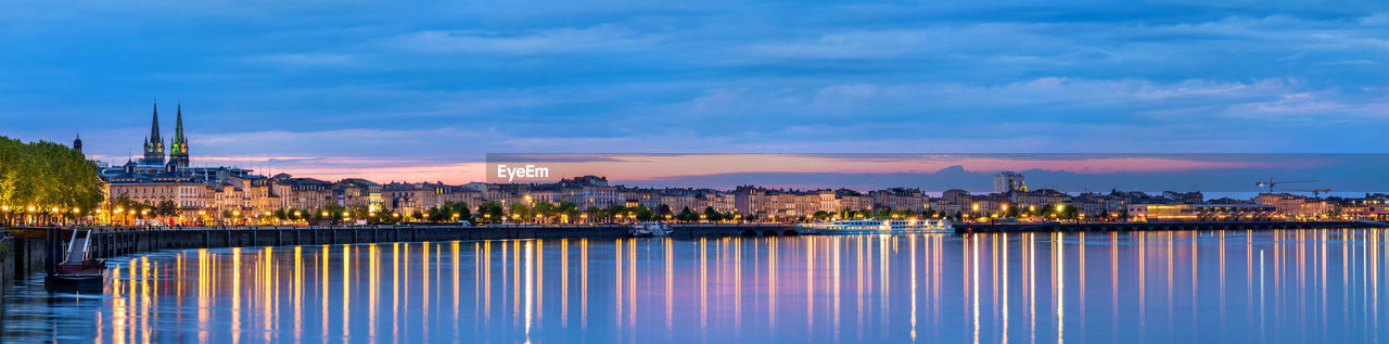 ILLUMINATED BUILDINGS BY RIVER AGAINST SKY AT DUSK