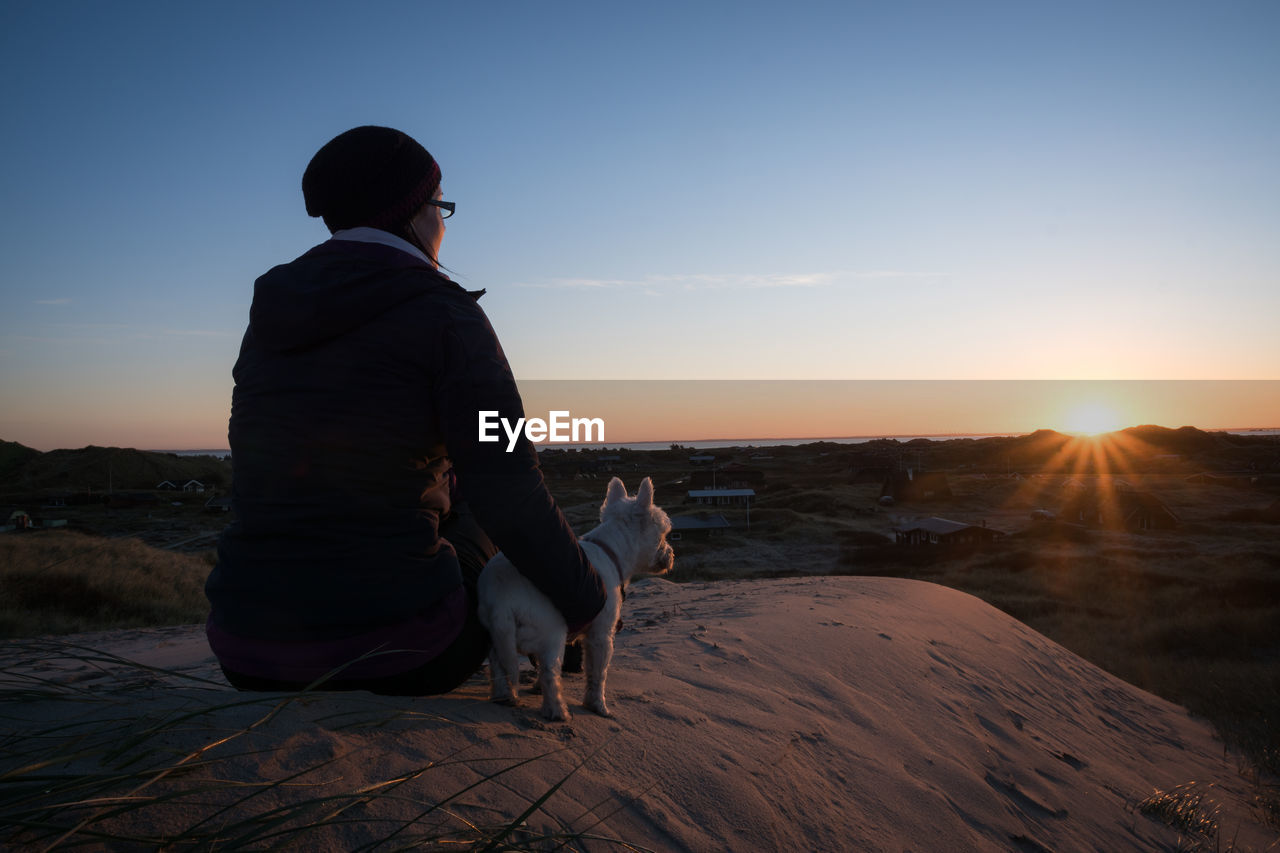 Rear view of woman with dog sitting on landscape at sunset