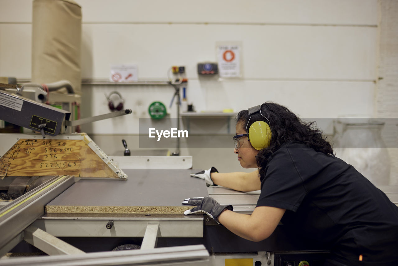 Side view of female carpenter wearing protective eyewear and ear protectors while working in factory