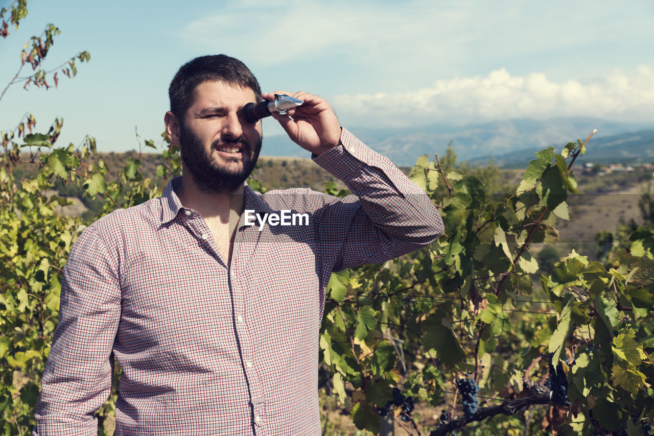 Man looking through refractometer while standing against plants and sky