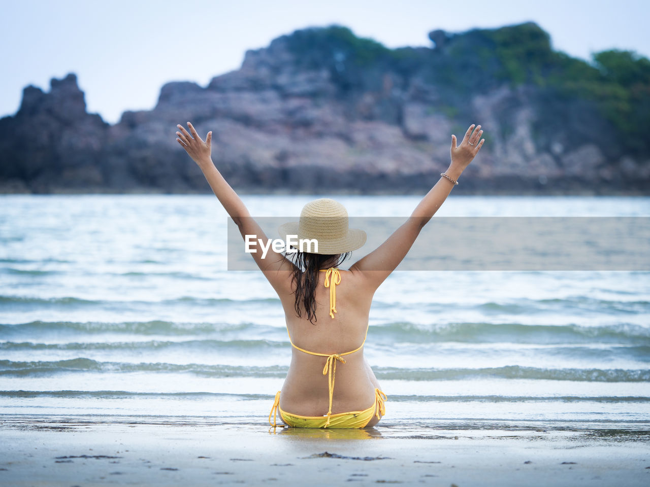 Rear view of woman wearing bikini with arms outstretched while sitting at beach
