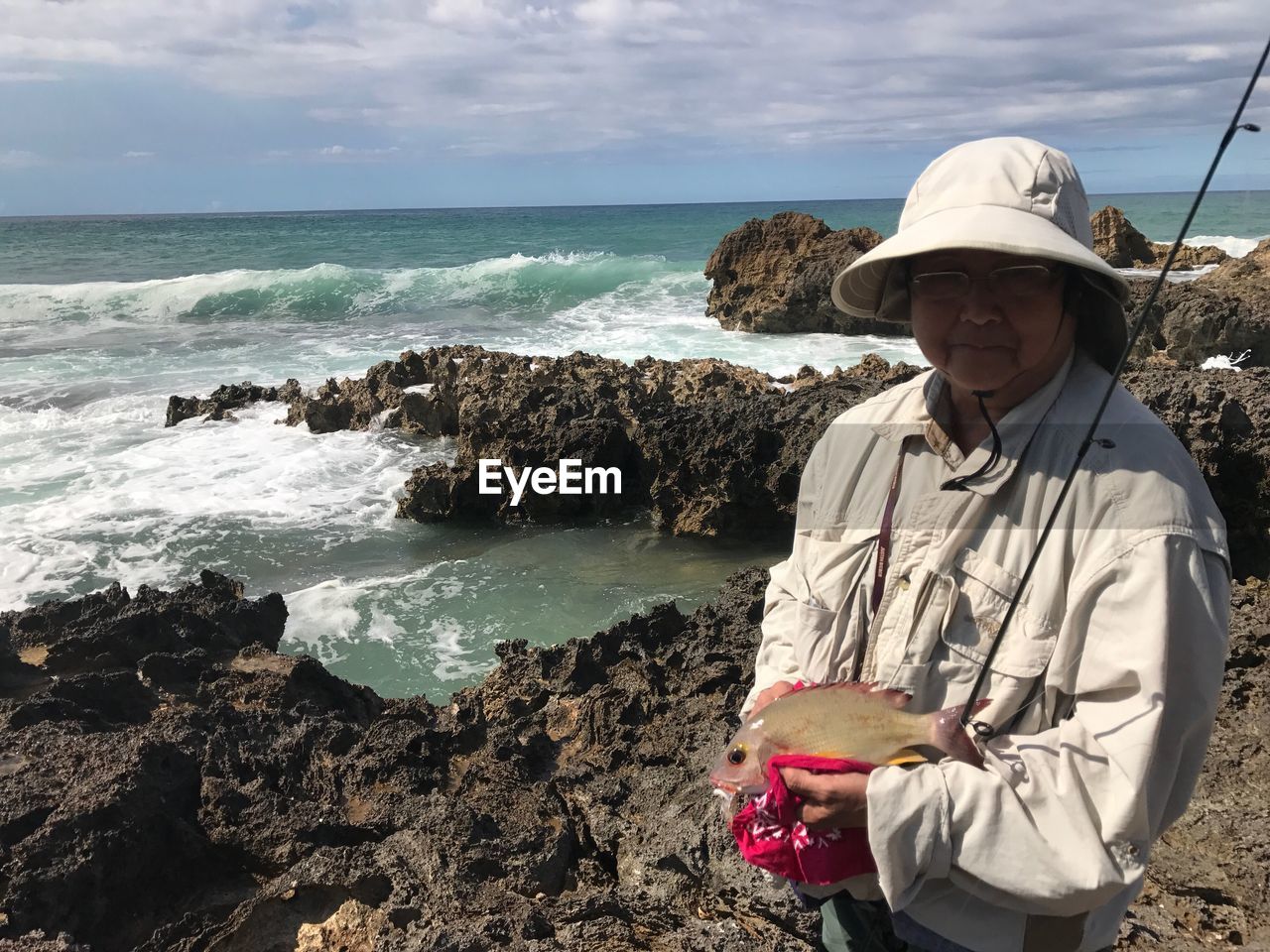 Portrait of senior woman holding fishing rod while standing at beach against sky