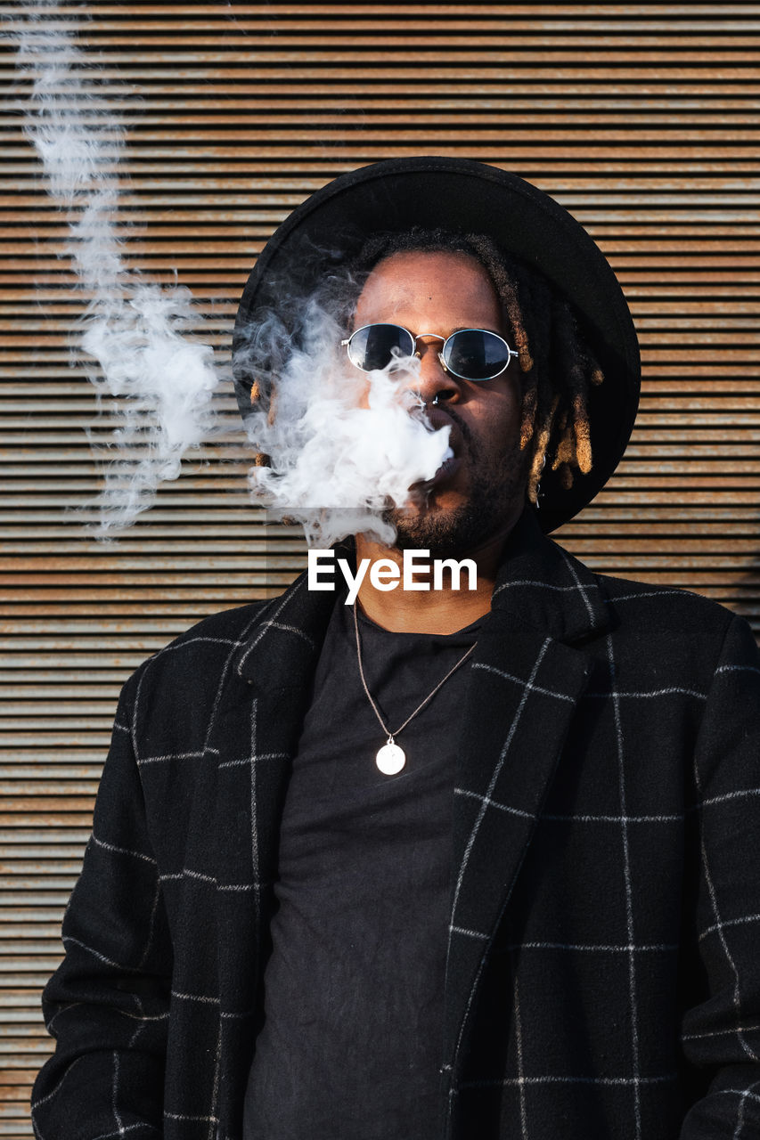 Modern african american man with dreadlocks wearing hat with sunglasses and exhaling smoke on street