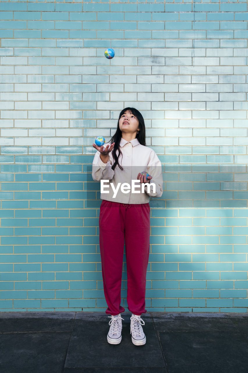 Young woman juggling with small globes in front of turquoise brick wall