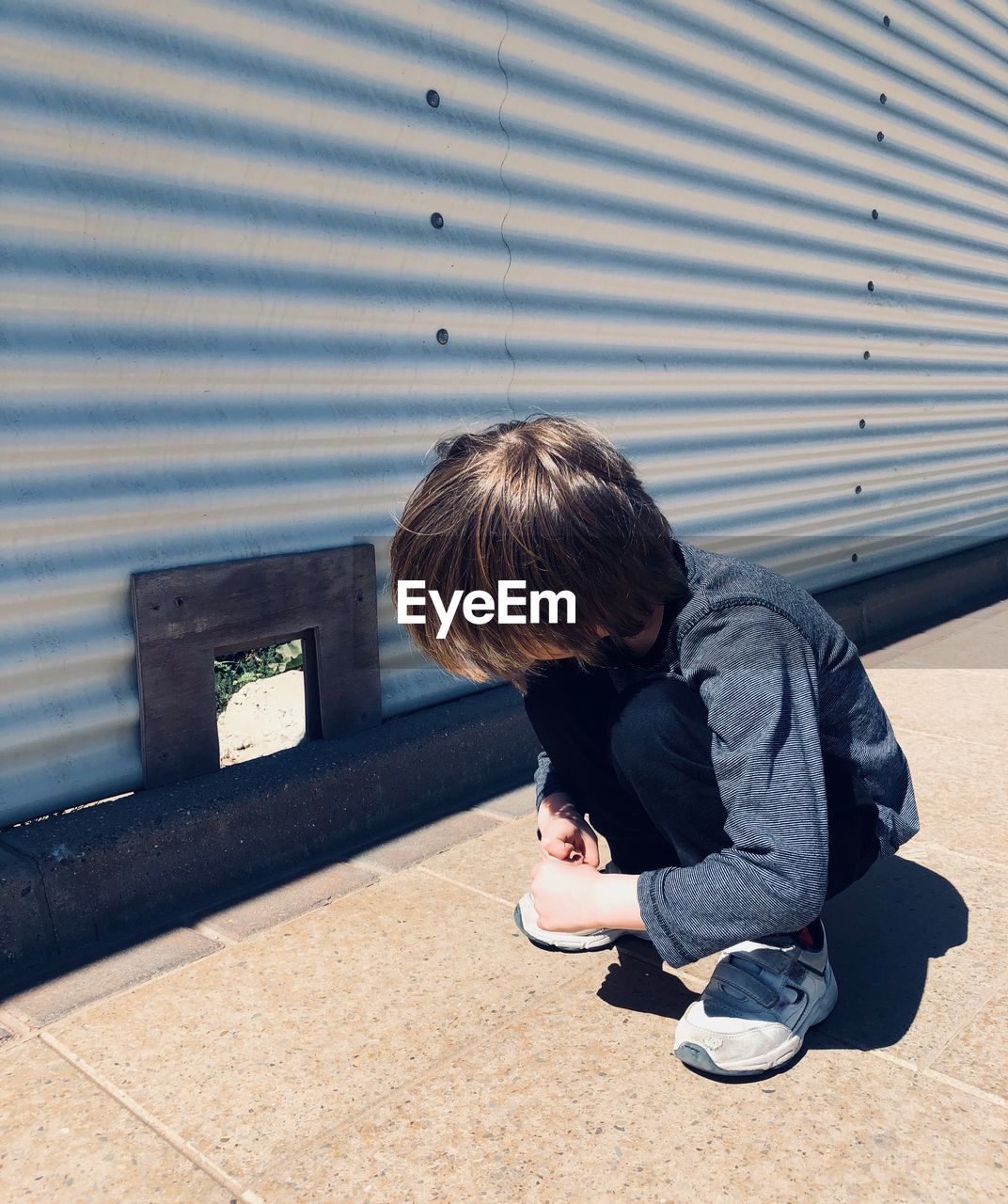Boy looking at corrugated iron while crouching on sidewalk during sunny day