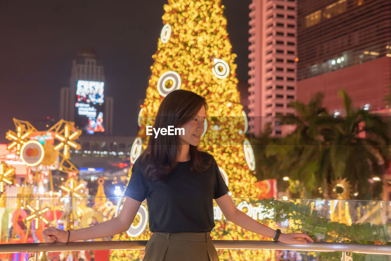 Smiling woman standing against illuminated christmas tree in city at night