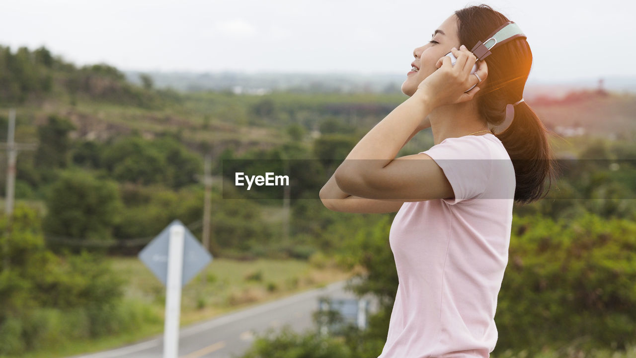 Side view of smiling young woman listening music against sky