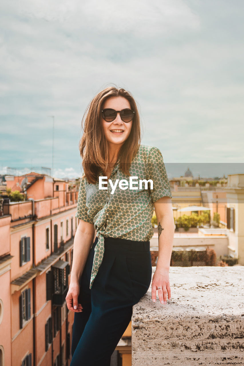 A young smiling woman in sunglasses sits on a rooftop with a panoramic view of rome, italy