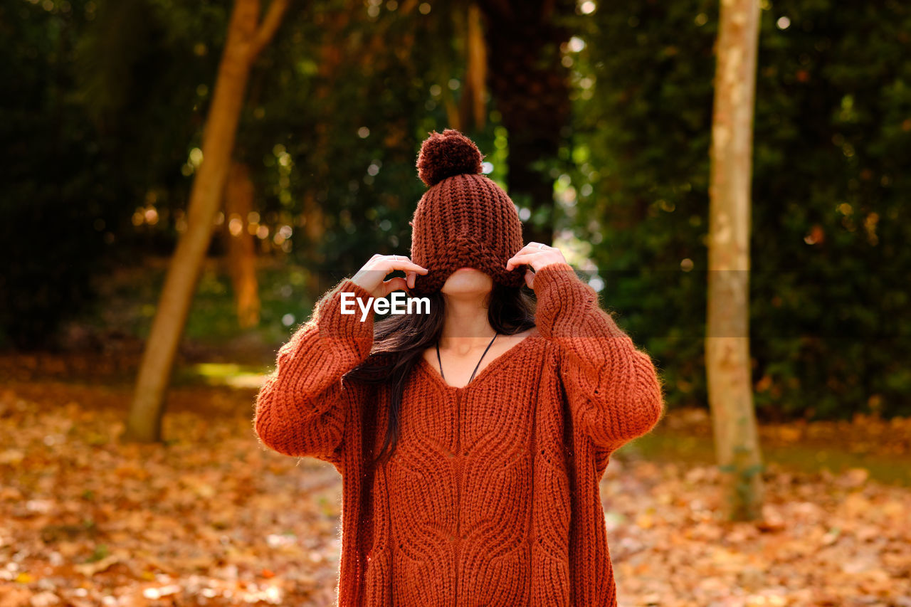 Unrecognizable female in knitted hat covering face wearing a warm sweater while standing in forest in cold autumn day