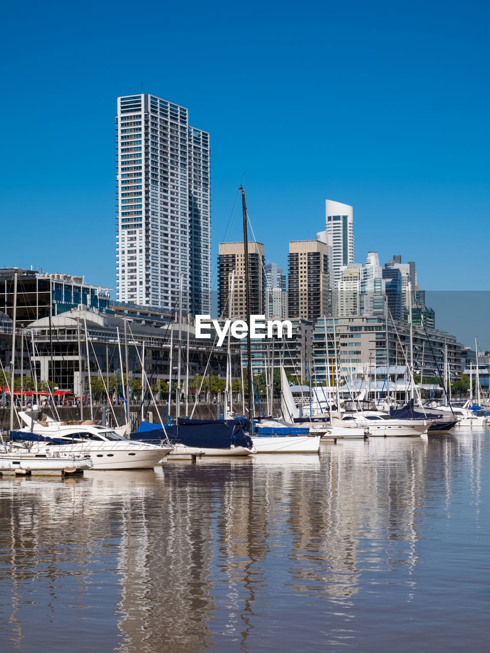 SAILBOATS MOORED AT HARBOR AGAINST BUILDINGS IN CITY