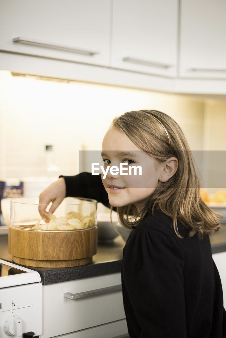 Side view portrait of girl eating potato chips at kitchen counter