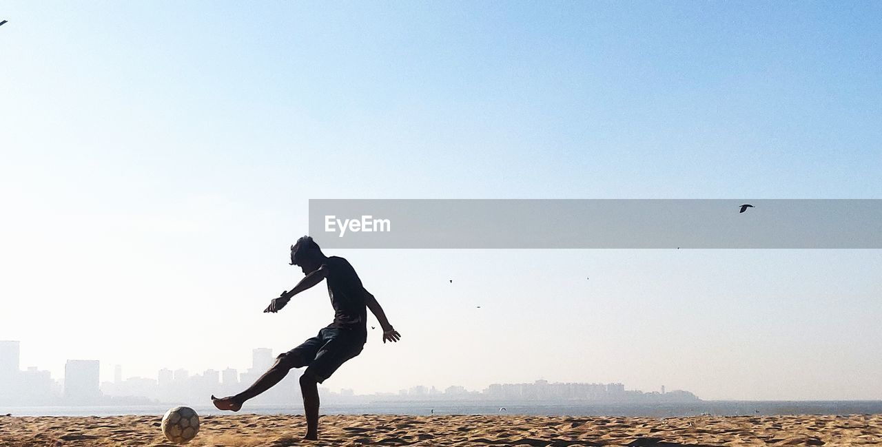 Teenage boy playing soccer at beach against clear sky