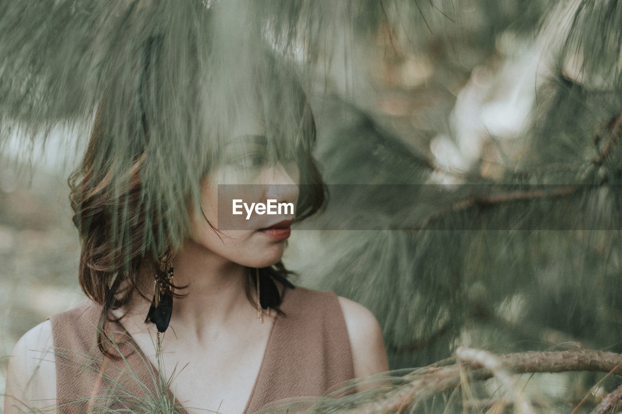 Close-up of thoughtful young woman looking away while standing by trees at park