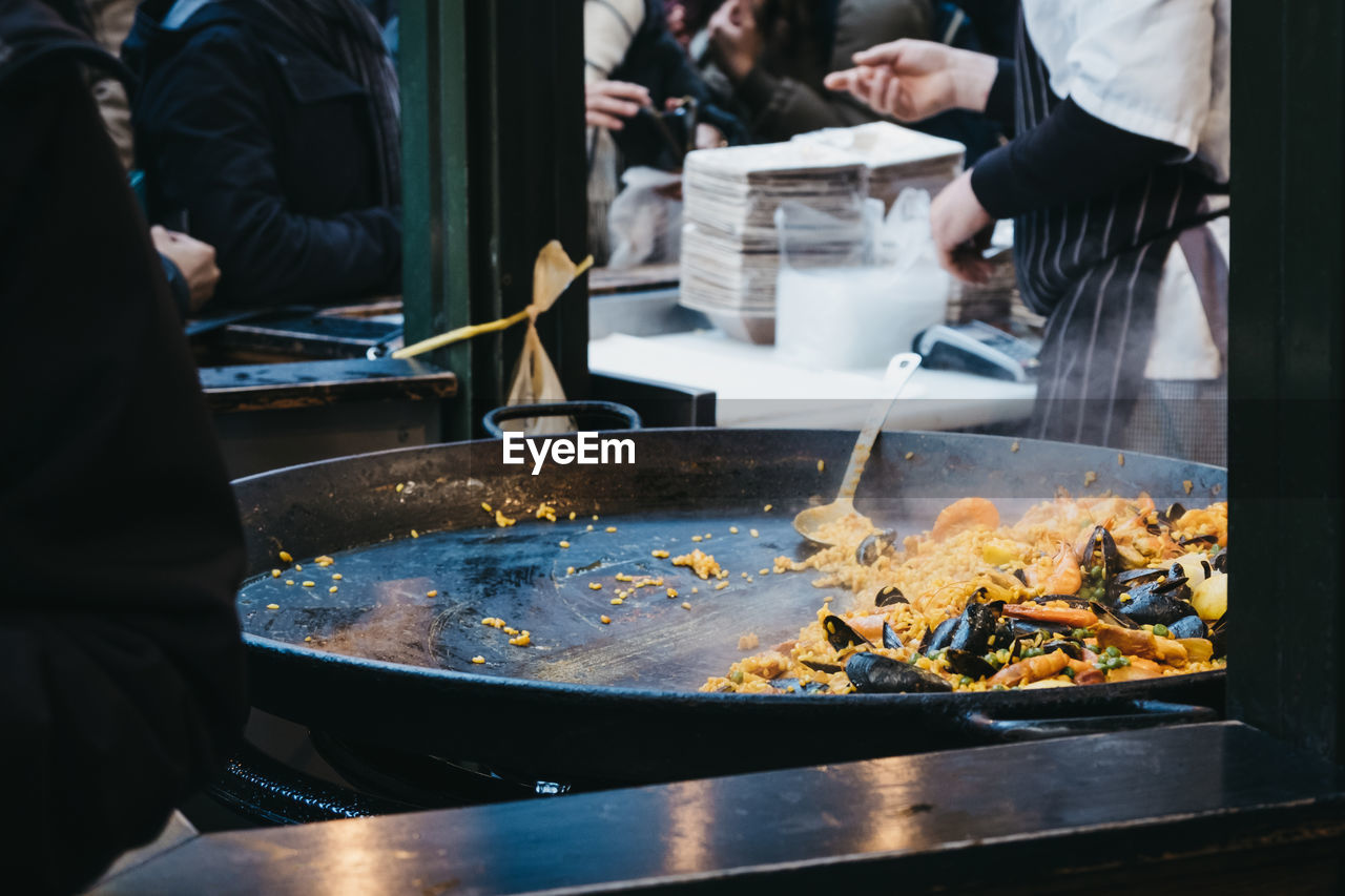 Large pan of paella at a street food market, unidentifiable people on the background.