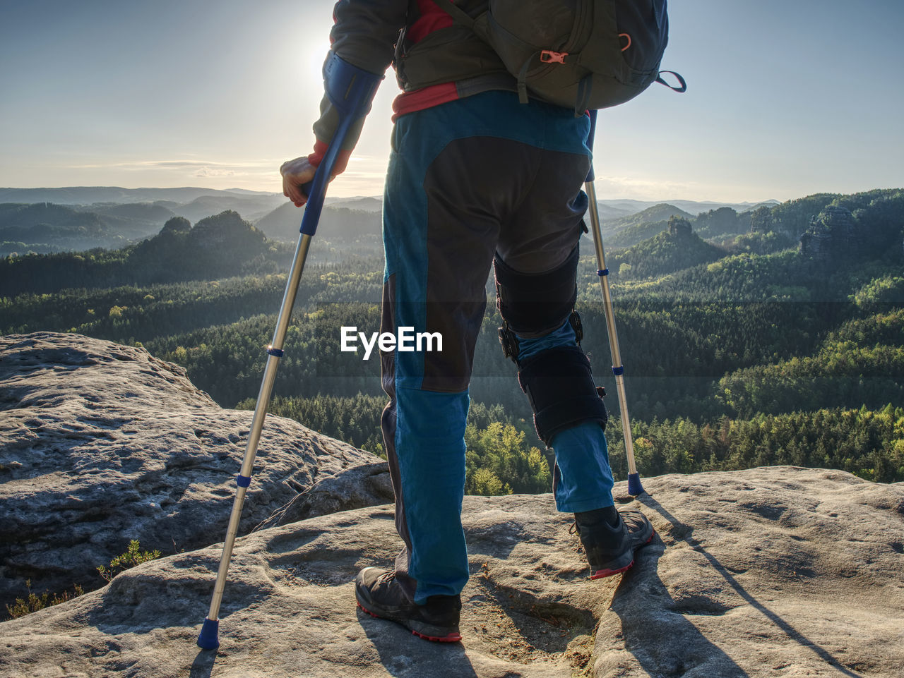 Happy man hiker hold medicine stick, injured knee fixed in knee brace feature. scenic mountain top 