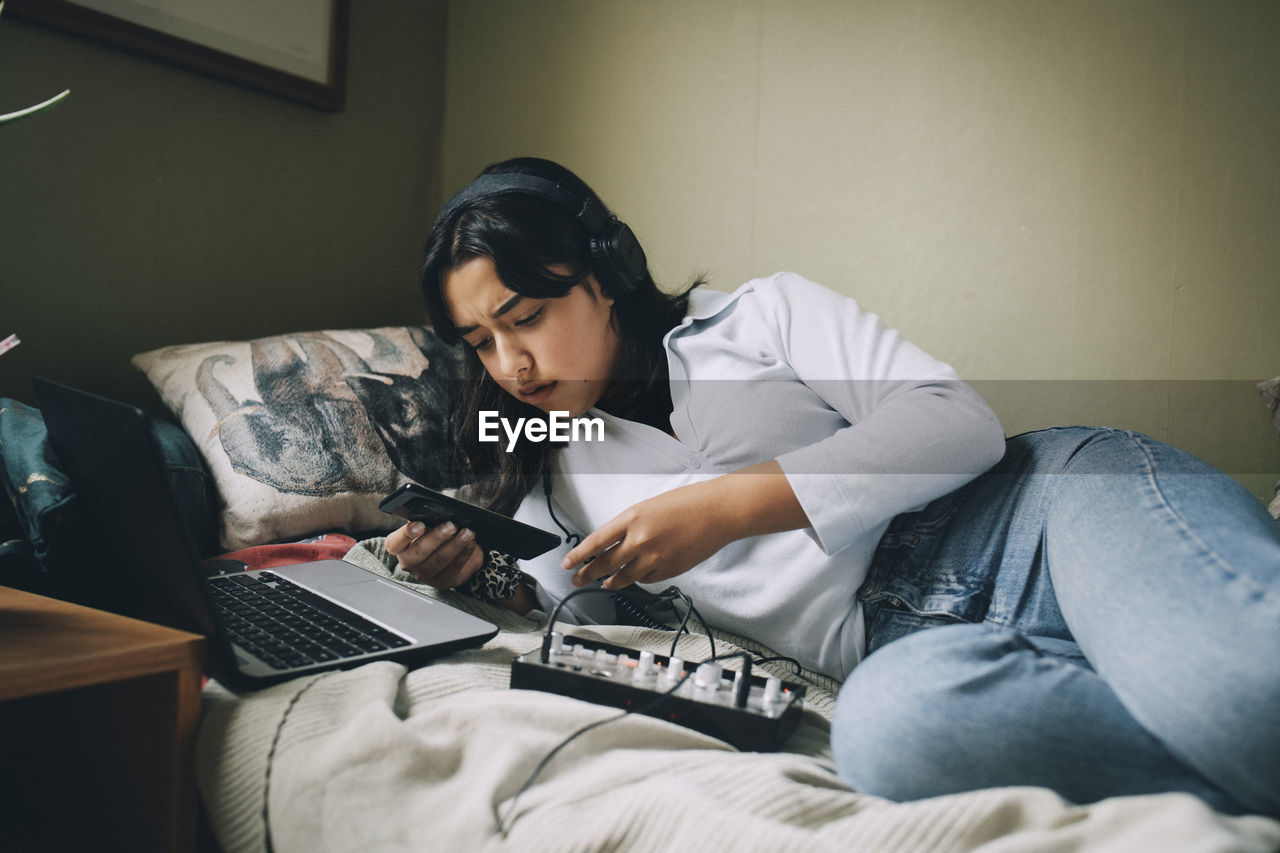 Teenage girl using mobile phone and laptop while lying on bed at home