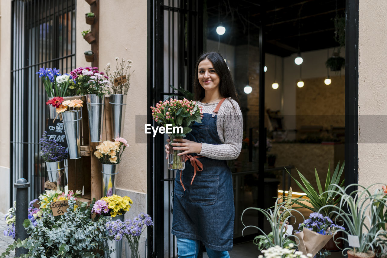 Young female florist holding flower vase while standing at shop