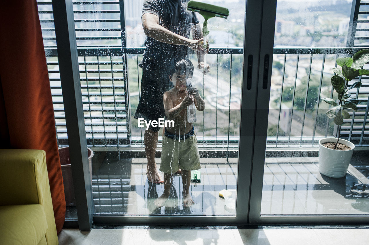 Low section of father with son cleaning windows in balcony seen through glass