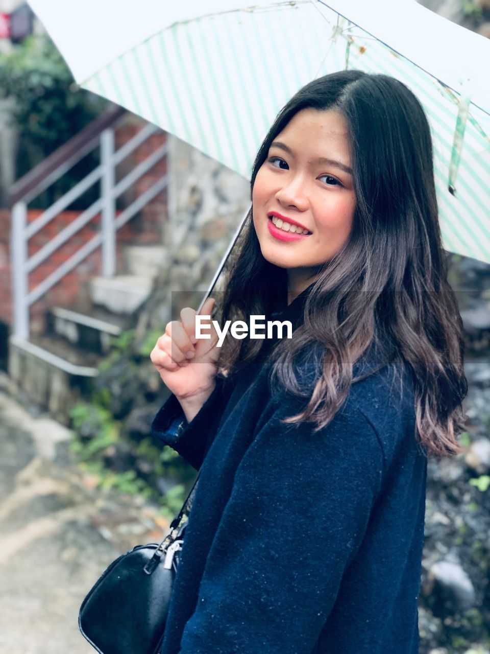 PORTRAIT OF SMILING YOUNG WOMAN STANDING AGAINST BLURRED BACKGROUND