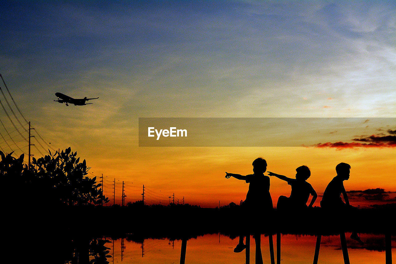 Silhouette boys pointing at airplane against sky during sunset