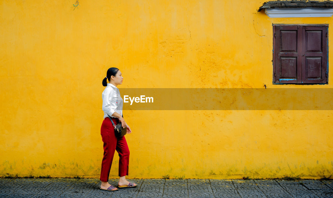 Side view of young woman standing on road against wall