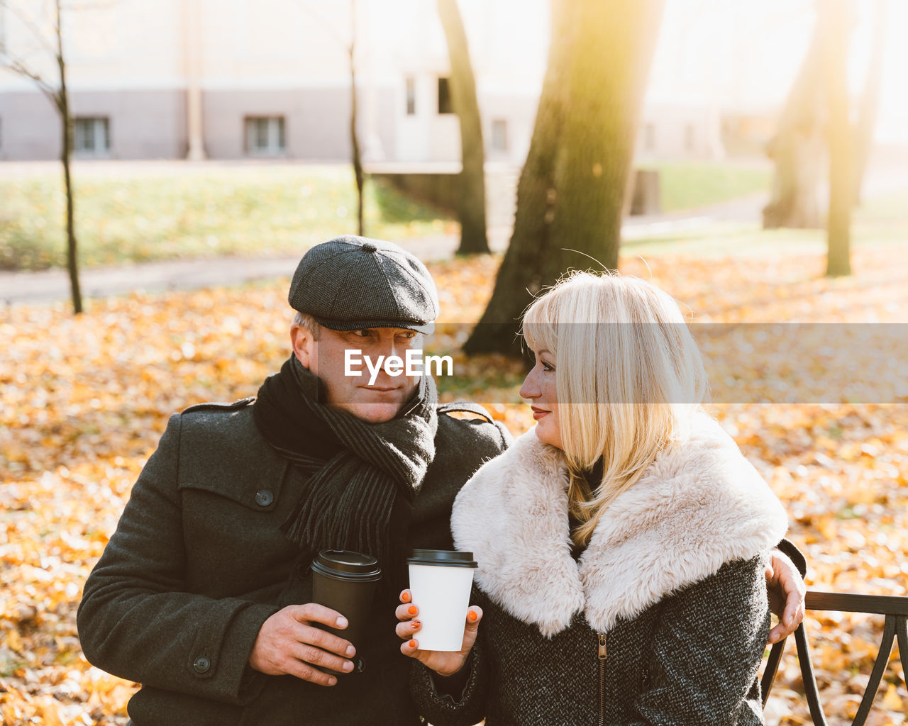 Mature couple holding disposable coffee cup while sitting on bench in park during autumn