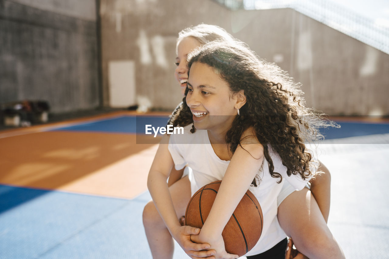 Smiling girl carrying piggyback to female friend at sports court