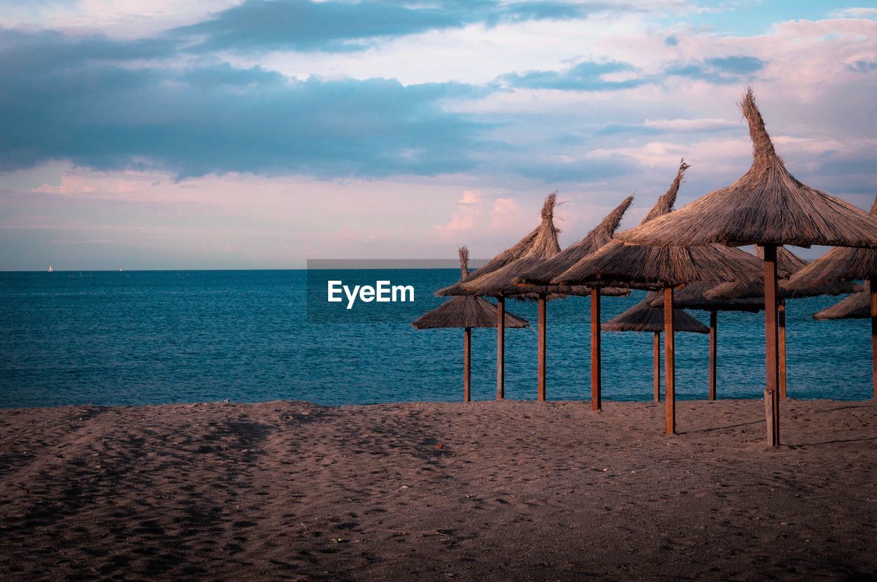 Thatched roofs on beach against cloudy sky during sunset