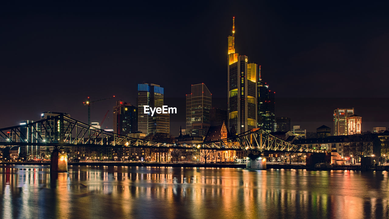 Illuminated bridge over river by buildings against sky at night