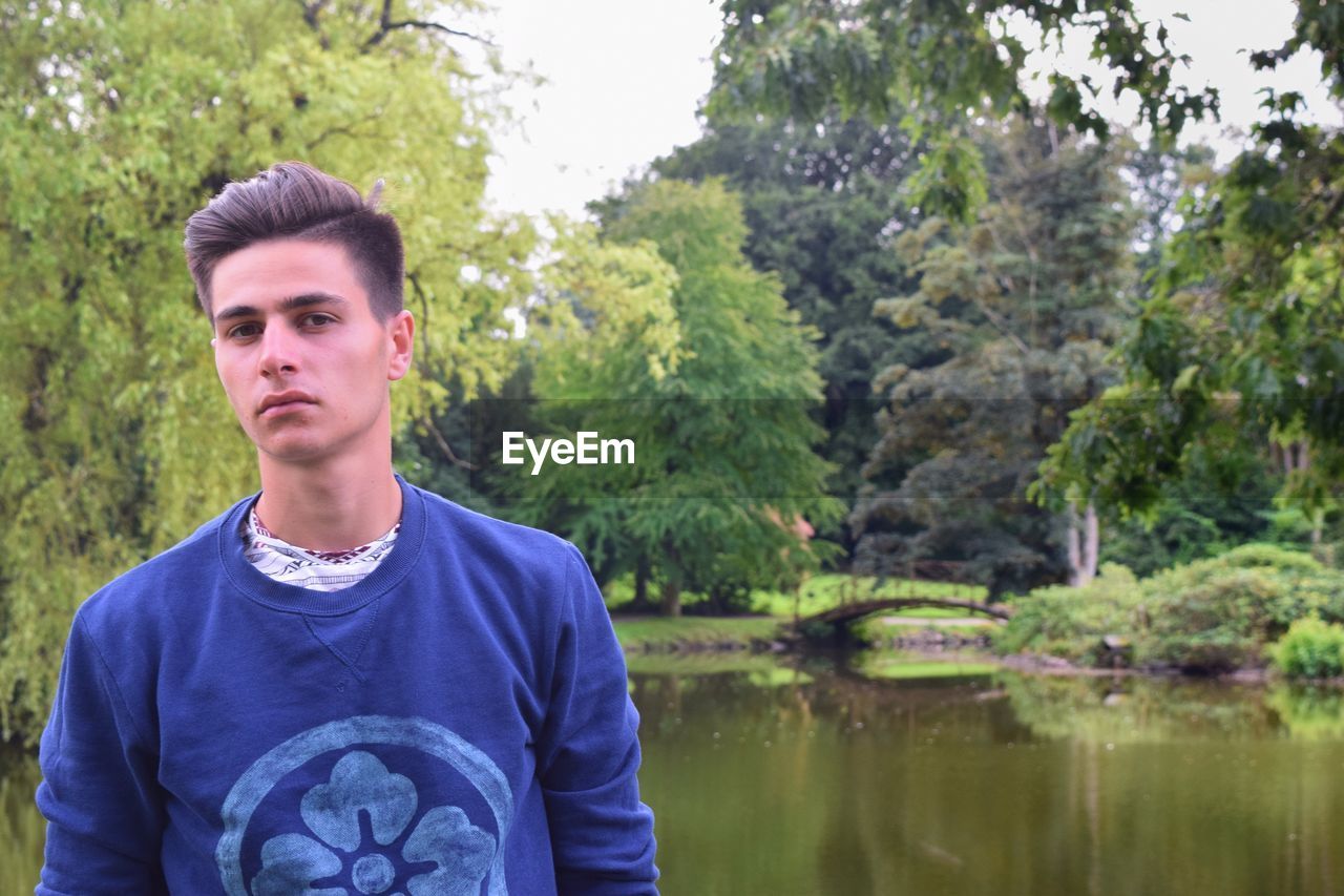 Portrait of young man standing by lake against trees