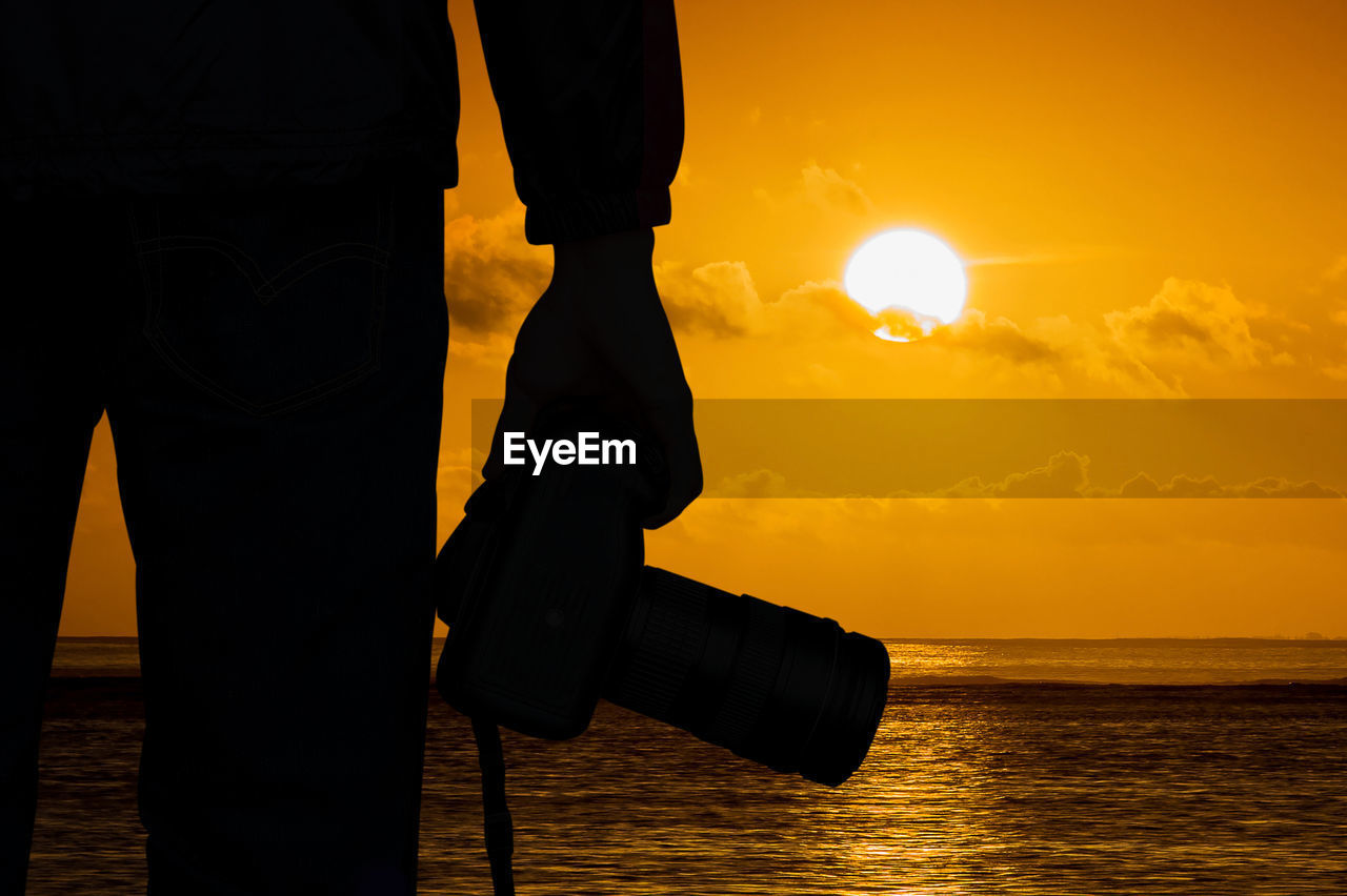 Close-up of silhouette man holding camera by sea during sunset