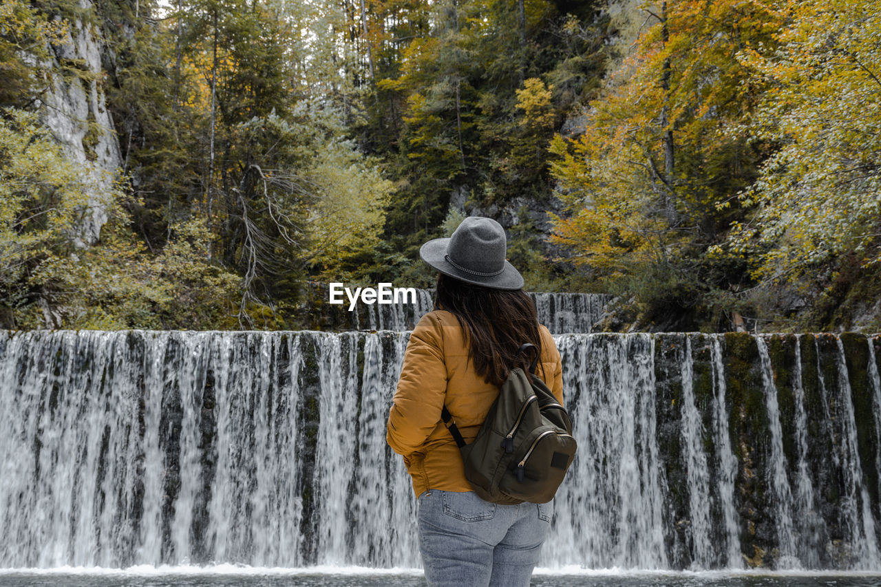 waterfall, one person, water, tree, motion, nature, leisure activity, clothing, beauty in nature, plant, rear view, flowing water, hat, scenics - nature, autumn, lifestyles, adult, water feature, winter, day, standing, forest, casual clothing, three quarter length, women, young adult, long exposure, outdoors, spring, vacation, trip, travel, land, backpack, activity, tourism, fashion accessory, holiday, non-urban scene, blurred motion, river, environment, warm clothing, men, splashing, hiking, person