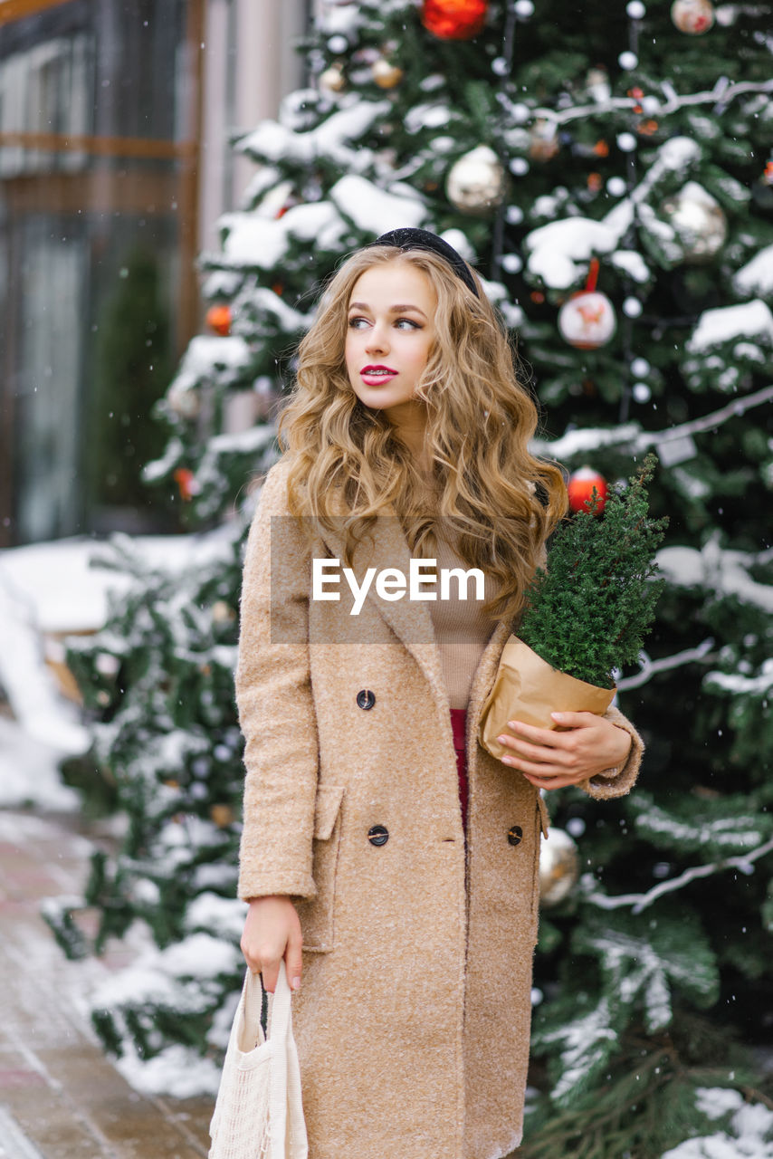 A pretty blonde holds a potted christmas tree in her hands, which she bought in a store, and walks