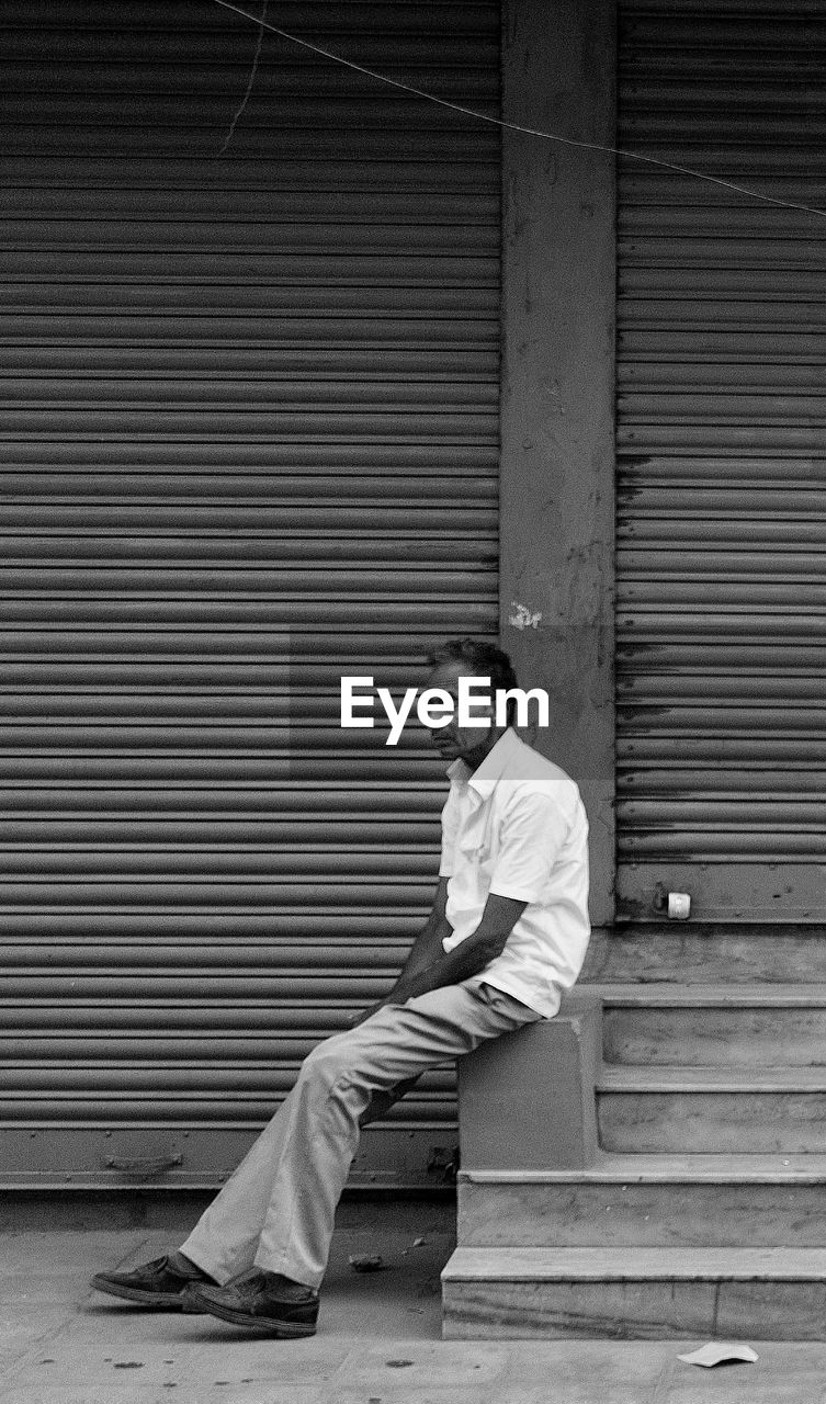 Side view of young man sitting against closed shutter
