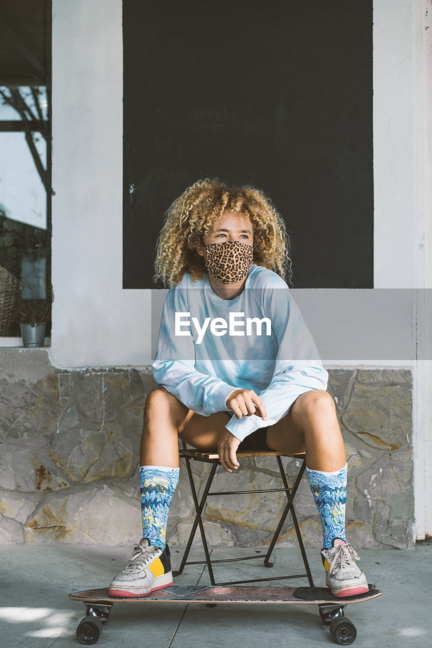 Woman wearing face mask sitting on chair with skateboard against wall