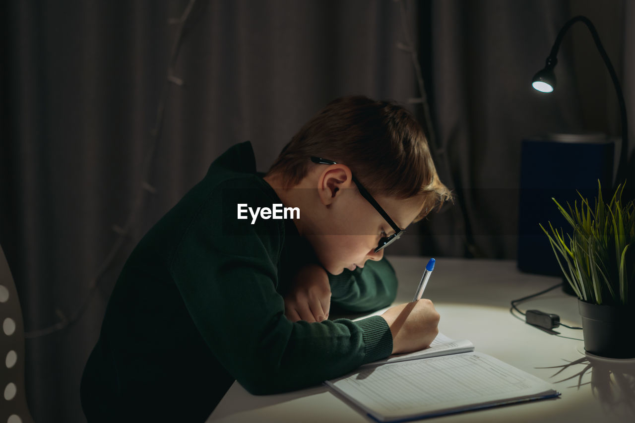 Caucasian boy doing homework at home in dark room by the light of lamp.