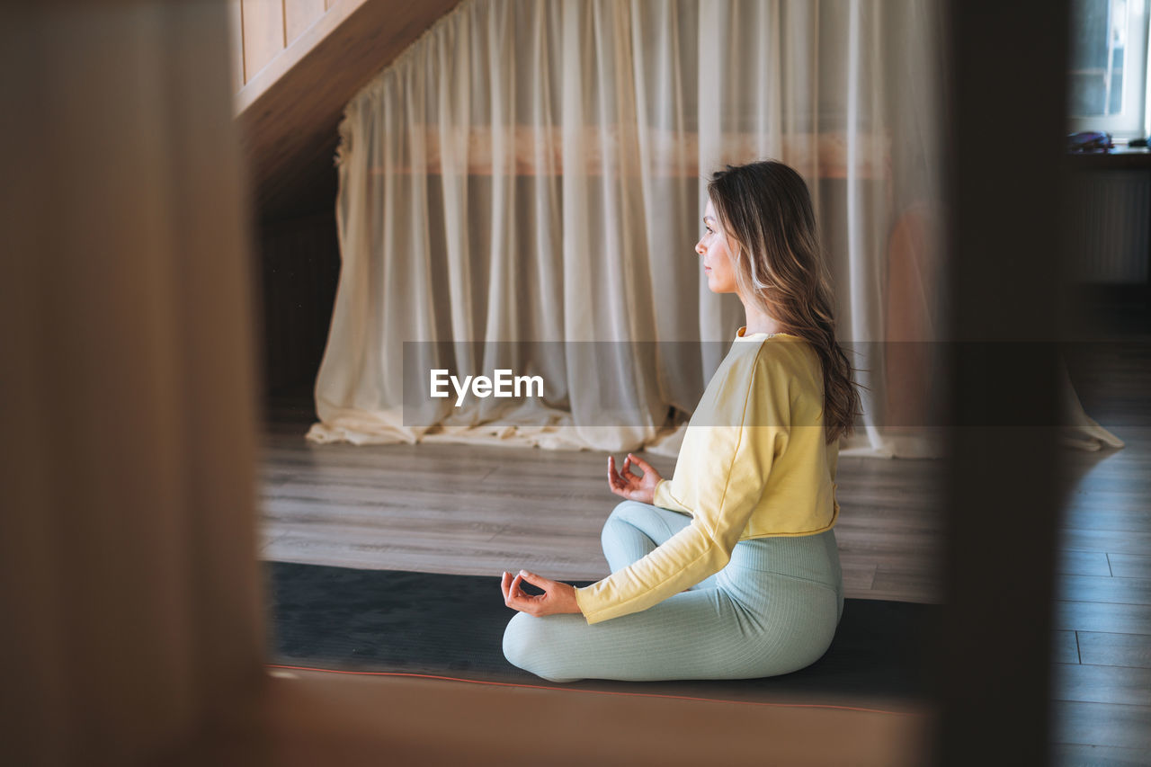 Young slim fitness blonde woman practice morning yoga near window of the home