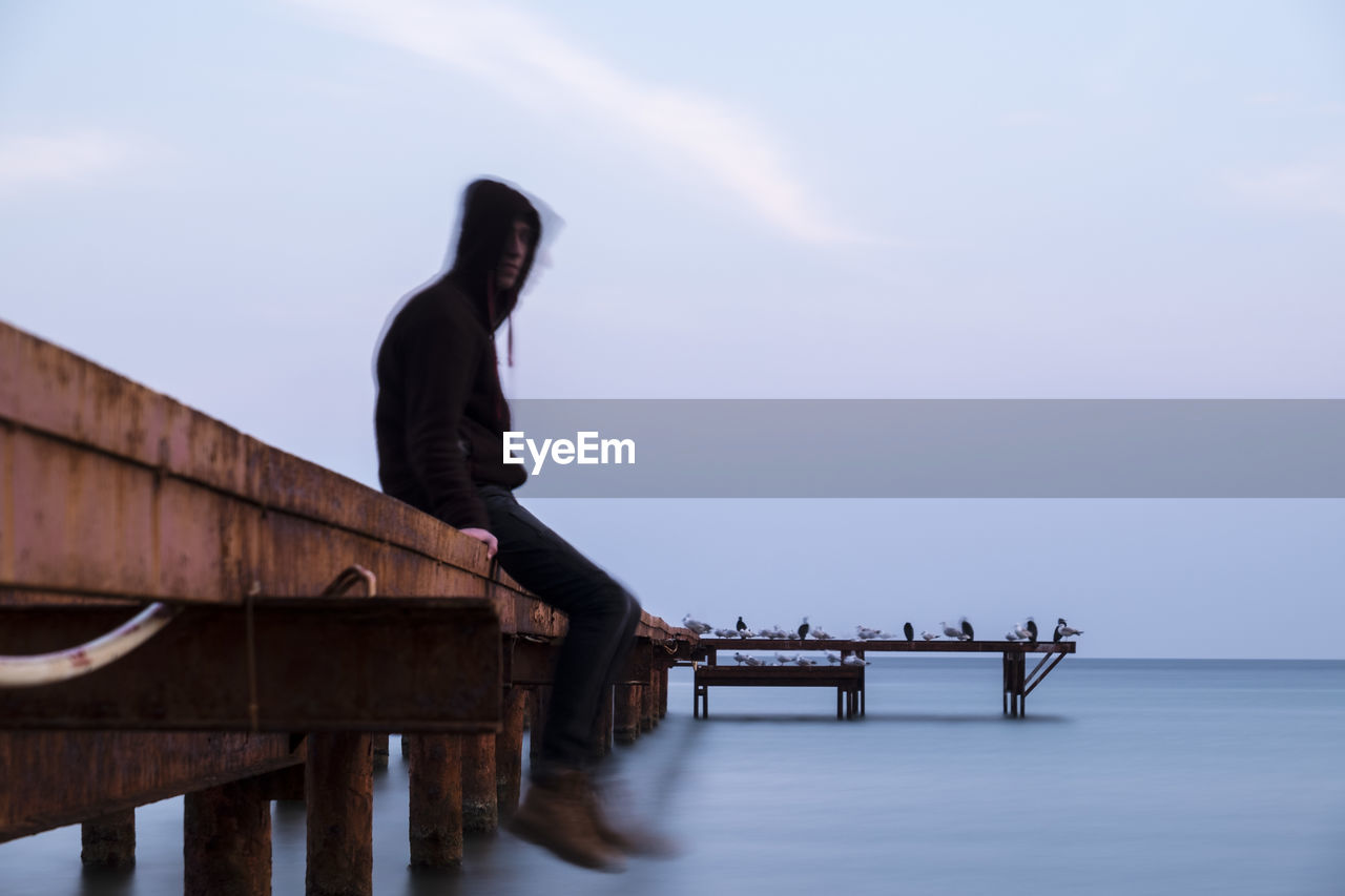 SIDE VIEW OF MAN SITTING ON PIER OVER SEA